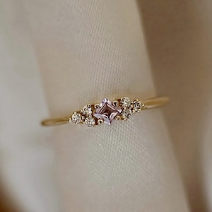 Könnte beinhalten: Ein zarter goldener Ring mit einem quadratischen, hellvioletten Edelstein in der Mitte, flankiert von Gruppen kleiner, klarer Edelsteine. Der Ring hat ein dünnes, goldfarbenes Band mit einem schlichten, eleganten Design. Der Ring ist vor einem weichen, cremefarbenen Hintergrund platziert.