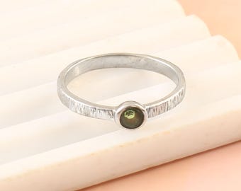 May include: A silver ring with a textured band and a small, round, olive-green gemstone set in the center. The ring is simple and elegant, suitable for everyday wear. The band has a subtle, hammered texture.