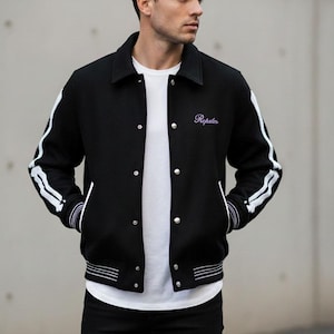 May include: Black varsity jacket with white stripes on the sleeves and a white trim. The jacket has a button-up front and a small embroidered word on the chest. The person in the image is wearing a white t-shirt and black pants.