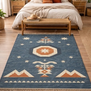 May include: Blue area rug with geometric patterns in orange and white. The rug features a central star design, mountain motifs, and fringe trim. The rug is placed in a bedroom setting with a wooden bed frame and nightstands.