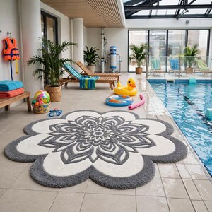 May include: A large, circular, flower-shaped rug featuring a central grey and white mandala pattern with scalloped edges. The rug is textured with a plush pile and displays a symmetrical floral design in shades of dark grey and off-white.