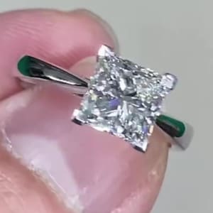 Puede incluir: Un anillo de plata con un diamante de talla cuadrada. El diamante es el punto focal, reflejando la luz. El anillo es sostenido por dedos, mostrando el diseño y el brillo. Es una pieza de joyería.