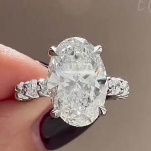 Pode incluir: Um close-up de um anel de diamante com uma pedra central de corte oval e uma banda adornada com diamantes menores. O anel é cravejado em um metal prateado. O texto "GOLD FLUSH" está na parte inferior.