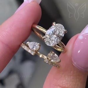 Puede incluir: Dos anillos de oro con detalles de diamantes. Uno presenta un gran diamante de talla ovalada, y el otro tiene dos diamantes en forma de pera y piedras de acento más pequeñas. Los anillos están sobre un fondo rosa suave. El texto "GOLD FLUSH" es visible.