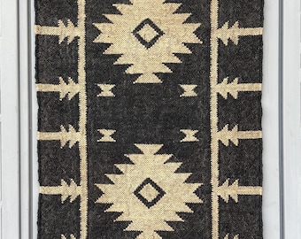 May include: A rectangular, dark gray rug with a geometric pattern in beige. The design features diamond shapes and zig-zag lines. The rug has a fringed edge at the bottom and is likely made of woven material.
