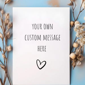 May include: A white card with the text "YOUR OWN CUSTOM MESSAGE HERE" and a heart symbol. The card is surrounded by dried floral arrangements against a light blue background.