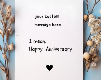 May include: White greeting card with the text "your custom message here" and "I mean, Happy Anniversary" in black font. A black heart is at the bottom. The card is on a light blue background with dried flowers.