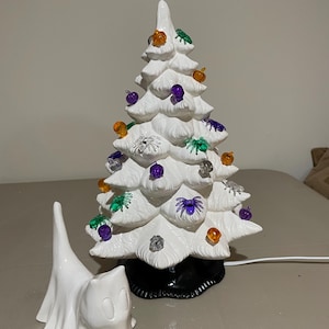 Op de afbeelding: Een witte keramische Halloween-boom versierd met oranje, paarse en groene ornamenten. Een klein wit spookfiguur staat voor de boom. De boom heeft een zwarte basis en een netsnoer.