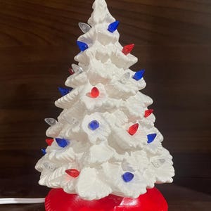 May include: A ceramic Christmas tree with a white finish, adorned with red, blue, and clear lights. The tree sits on a vibrant red base, creating a festive holiday decoration.