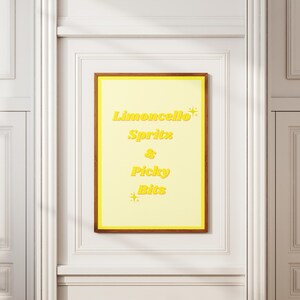 May include: A framed poster with the words "Limoncello Spritz & Picky Bits" in yellow text on a light yellow background. The frame is brown, and the poster is mounted on a white wall.