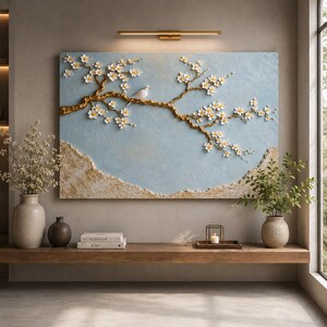 May include: A rectangular wall art piece featuring a textured light blue background with a raised, three-dimensional gold branch extending across the center. White cherry blossoms adorn the branch, and a small white bird sits perched on it.