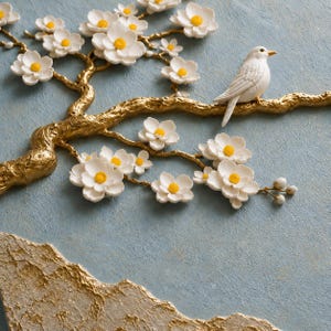 May include: A three-dimensional wall art piece featuring a textured gold branch extending across a light blue background. White flowers with bright yellow centers are attached to the branch, and a white bird is perched on the golden limb.