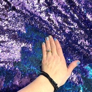 May include: A hand resting on a fabric with iridescent purple and blue sequins. The sequins are arranged in a scale-like pattern.