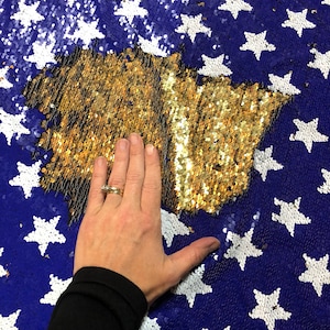 May include: A blue sequin fabric with white stars and a gold sequin area. The gold area is in the shape of a map. The fabric is being touched by a hand.