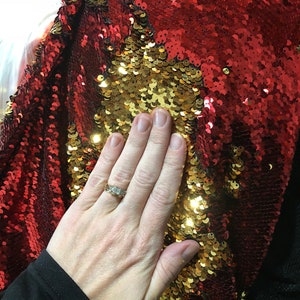 May include: Red and gold sequin fabric with a hand resting on the fabric. The sequins are arranged in a pattern that creates a mermaid tail effect.