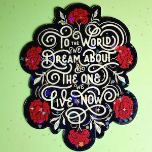 May include: A black sticker with white script reading "To the world we dream about & the one we live in now." The sticker features red floral accents and a holographic sheen.