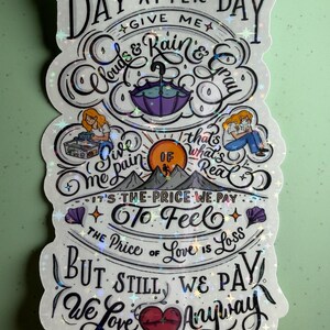 May include: A holographic sticker with the words "Day After Day" and other phrases like "Clouds & Rain & Gray" and "We Love Anyway." The sticker features illustrations of an umbrella, mountains, and a heart.