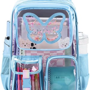 Puede incluir: Una mochila azul claro con un panel frontal transparente y un diseño en forma de mariposa. La mochila tiene múltiples compartimentos, incluyendo bolsillos laterales y un bolsillo frontal con bolígrafos y rotuladores. Dentro del panel transparente hay libros, un teléfono y un cuaderno.