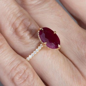 以下が含まれることがあります： 大きなオーバルカットのルビーの宝石をあしらったゴールドリング。ルビーは深い赤色で、小さな透明な宝石のハローに囲まれています。リングのバンドにも、小さな透明な宝石が飾られています。