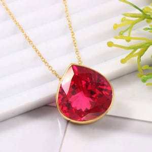 May include: A gold chain necklace featuring a large, faceted, pear-shaped red gemstone pendant set in a gold bezel. The vibrant red stone has a brilliant cut that reflects light, suspended from a delicate gold link chain that secures the pendant.