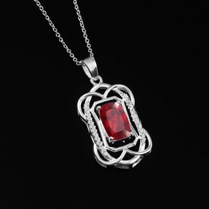 May include: A silver-toned pendant necklace featuring a rectangular, faceted deep red gemstone at the center. The stone is framed by an intricate, openwork silver metal design accented with small, clear stones along the side edges.