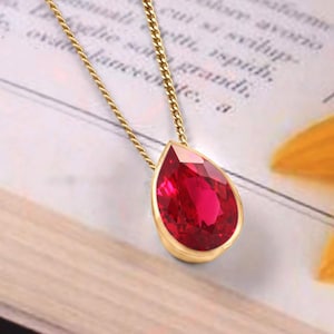 Peut inclure: Un collier à chaîne dorée doté d'un pendentif en forme de goutte avec une pierre précieuse rouge vif. Le pendentif est serti dans une monture dorée lisse, et la chaîne se compose de petits maillons dorés entrelacés partant du haut du sertissage.
