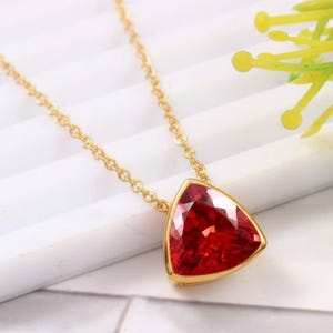May include: A gold-toned necklace featuring a triangular-cut, deep red gemstone pendant set in a smooth metal bezel. The pendant is suspended from a delicate, matching gold-toned chain with small, uniform circular links that catch the light.