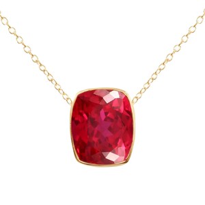 May include: A necklace featuring a cushion-cut, deep red gemstone set in a gold bezel frame. The pendant is suspended from a delicate gold chain with small, round links. The gemstone displays a faceted surface that reflects light.
