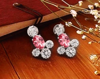 May include: A pair of silver earrings featuring a cluster design. Each earring has a central oval pink gemstone, surrounded by smaller, round, clear gemstones. The earrings are set against a warm, wooden background.