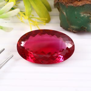 Könnte beinhalten: Ein facettierter, oval geformter Edelstein in leuchtendem Rot. Der Edelstein zeigt ein komplexes Schliffmuster, das Licht reflektiert. Das Bild zeigt auch ein grünes Mineral und gelbe und weiße florale Elemente.
