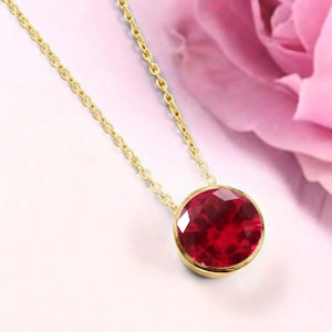 May include: A gold chain necklace featuring a round, faceted, deep red gemstone pendant set in a smooth gold bezel. The pendant hangs from a delicate gold link chain, showcasing a brilliant cut and reflective surface on the circular red stone.