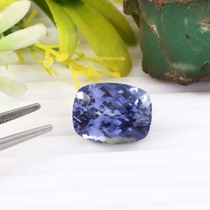 Op de afbeelding: Een gefacetteerde, kussenvormige, blauwe edelsteen. De edelsteen heeft een complex patroon van interne reflecties, met een mix van lichte en donkere blauwtinten. De edelsteen staat tegen een witte achtergrond, met een pincet en wat groen op de achtergrond.