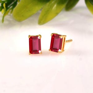 May include: A pair of stud earrings featuring rectangular emerald-cut red gemstones set in gold metal prongs. The earrings have a classic post-back design, showcasing the vibrant red color of the stones against the polished gold setting.