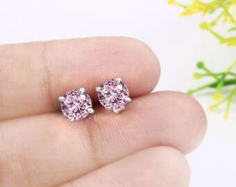 May include: A pair of stud earrings featuring round, light pink gemstones set in a silver metal. The earrings are displayed on a person's hand, with a soft background of white and yellow flowers.