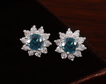 May include: A pair of stud earrings with a central teal gemstone, encircled by smaller, clear stones. The earrings have a floral design and are set in a silver-coloured metal. The earrings are presented against a dark backdrop, showcasing their sparkle.