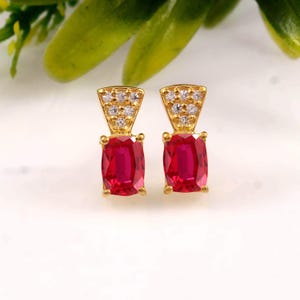 May include: A pair of gold stud earrings featuring a rectangular-cut vibrant red gemstone at the base. Above each red stone is a triangular gold setting embellished with six small, clear, sparkling white stones arranged in a cluster.