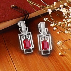 May include: A pair of silver-toned Art Deco style earrings. Each earring features a rectangular, deep red gemstone set within a geometric frame. Small clear stones are set above the red gems. The earrings are displayed on a wooden surface.