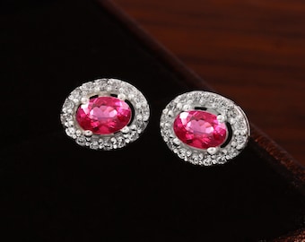 May include: A pair of stud earrings featuring oval-cut, bright pink gemstones surrounded by a halo of small, clear stones. The earrings are set in a silver-colored metal and are displayed against a dark background.