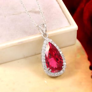May include: A silver necklace featuring a pear-shaped, deep red gemstone pendant surrounded by a halo of small, clear, sparkling stones. The pendant is suspended from a delicate silver chain, creating a classic and elegant jewelry piece.