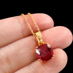 May include: A gold-colored pendant necklace featuring a large, square-cut, deep red gemstone. The pendant is set in a gold frame and adorned with small, clear accent stones. The necklace chain is also gold-colored.