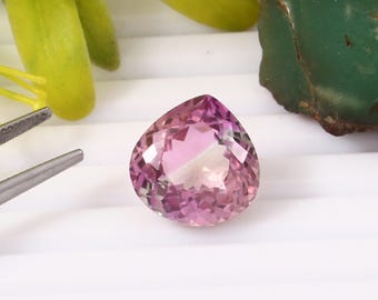 May include: A faceted, pear-shaped gemstone with shades of pink and purple. The gem is set against a white background, with a pair of tweezers and green and yellow organic elements in the background. The gemstone is a focal point.