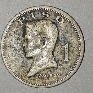 May include: A silver coin with the word "PISO" at the top and the number "1" on the right. The coin features a profile of a man and the text "JOSE RIZAL" at the bottom. The coin has a textured edge.