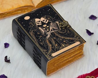 Op de afbeelding: Een zwart, leren notitieboek met gouden details en een sluiting. De omslag toont een hemels ontwerp met twee omarmende skeletten, met de woorden "The Lovers". De verouderde pagina's hebben een gouden tint.