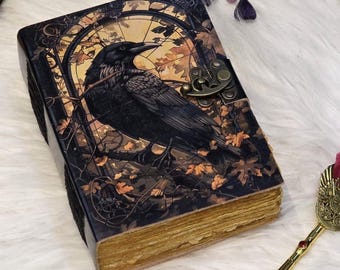 Può includere: Un diario rilegato in pelle scura con un'illustrazione di un corvo sulla copertina. Il corvo è appollaiato su un ramo contro uno sfondo di foglie autunnali e un quadrante dell'orologio. Il diario ha una chiusura in metallo e bordi delle pagine dorati.