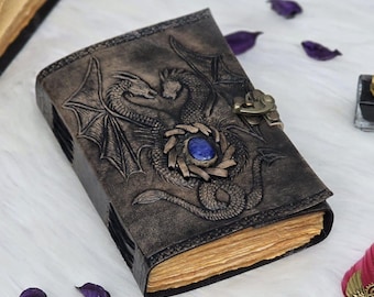 May include: A handcrafted leather journal with a detailed embossed dragon design. The cover features a central blue stone and a brass clasp. The pages have a vintage, aged appearance, suggesting a rustic or fantasy theme.