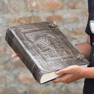 May include: A brown leather journal with a tree of life embossed on the cover. The journal has a clasp closure and stitched binding. The cover is a dark brown color with a detailed embossed design. The pages are white and the journal is held in the hands.