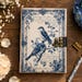 May include: A rectangular journal with a cream-colored cover featuring a blue floral and bird illustration. The design includes two birds perched on branches, surrounded by a decorative border and script text. A brass-colored metal latch secures the book.