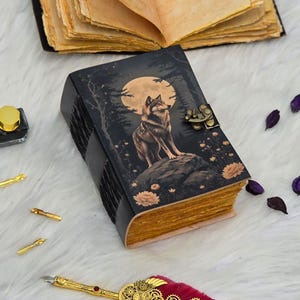 Peut inclure: Un journal en cuir noir avec une illustration de loup. La couverture présente un loup sur un rocher devant une pleine lune. Les pages ont un aspect vieilli et le journal est fermé par un fermoir en métal. Mesure environ 15 cm de long.