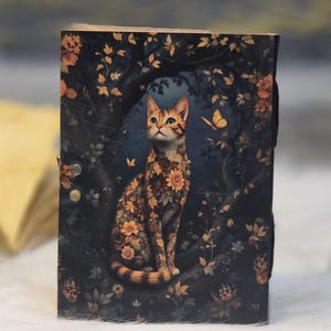 May include: A wooden journal with a cat illustration. The cat is covered in orange and yellow flowers, sitting on a tree branch against a dark blue background with leaves and butterflies. The journal has a black strap closure.