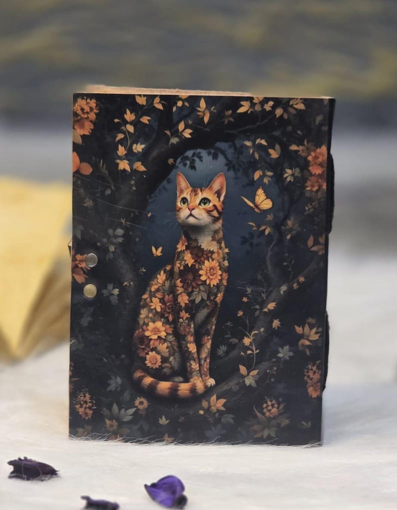 May include: A wooden journal with a cat illustration. The cat is covered in orange and yellow flowers, sitting on a tree branch against a dark blue background with leaves and butterflies. The journal has a black strap closure.